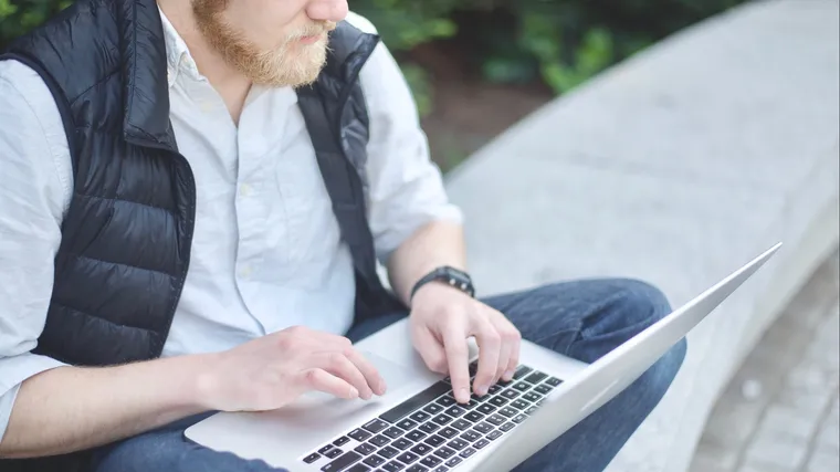 Picture of man typing on laptop while sitting on cement curb.